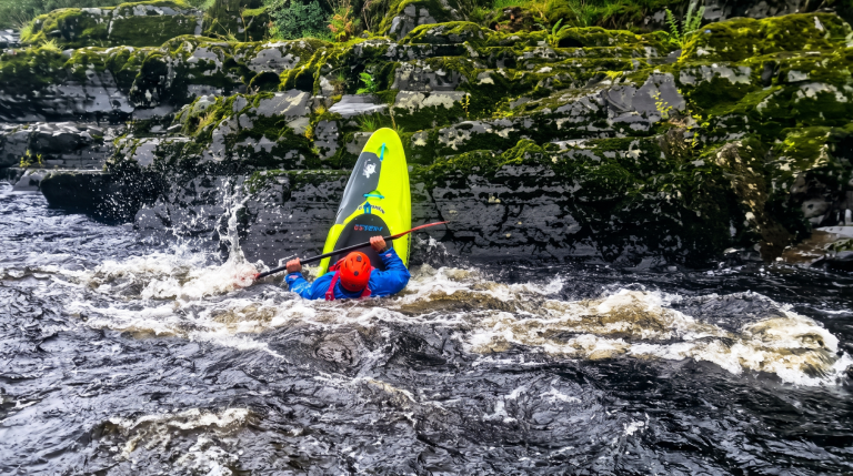 Kayaker in red helmet capsized in rapid river near mossy rocks.