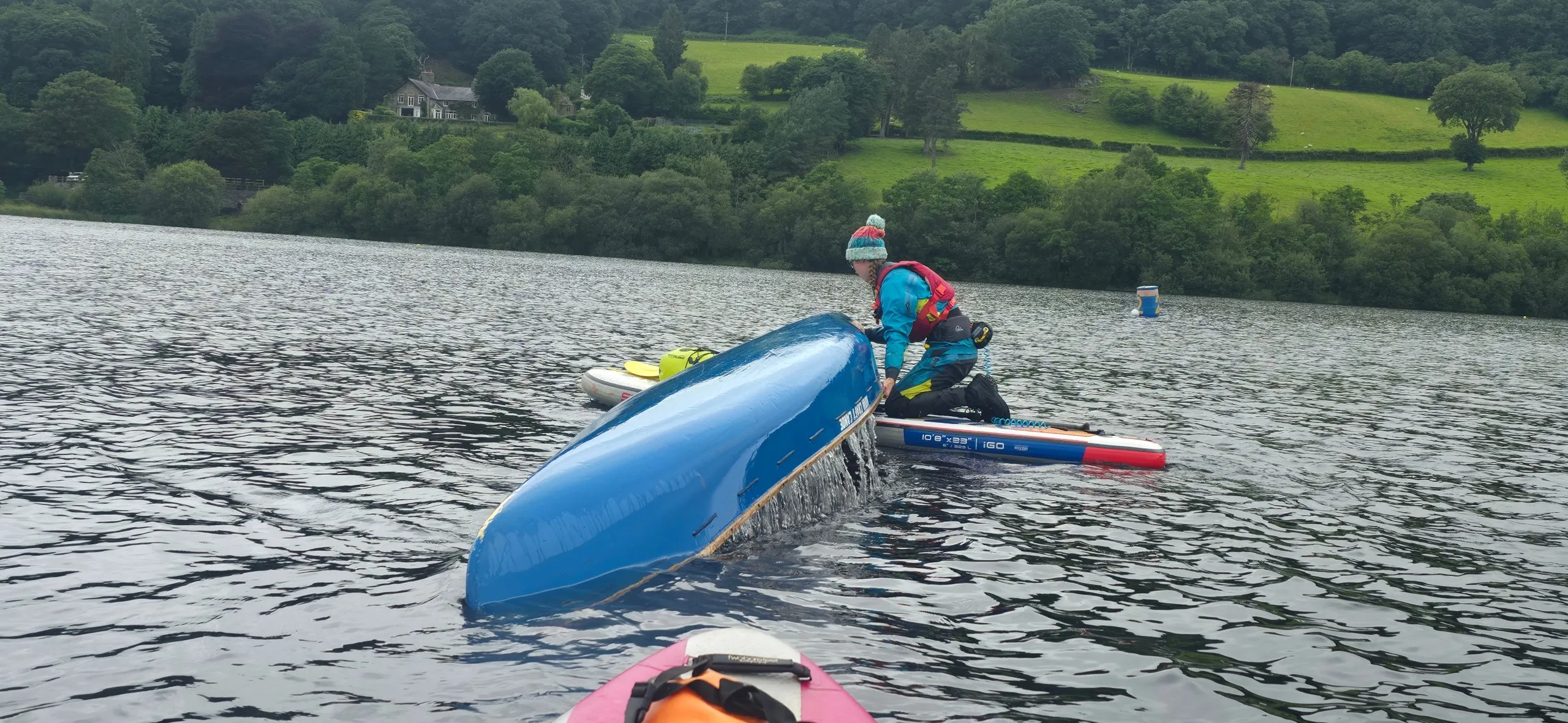 Person flipping blue kayak on a lake with green hills in the background.