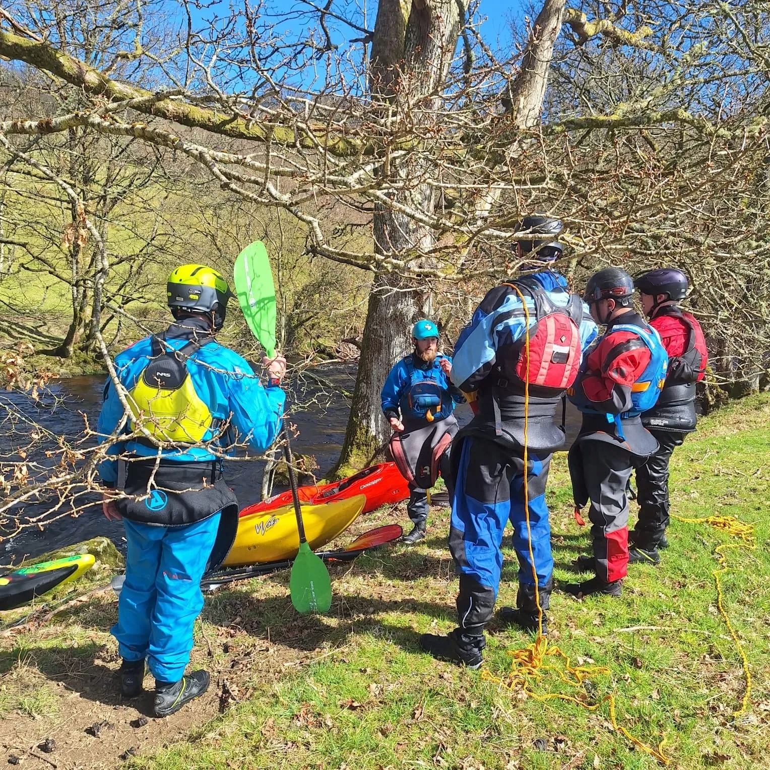 People in kayaking gear standing by a river with kayaks on a grassy bank.
