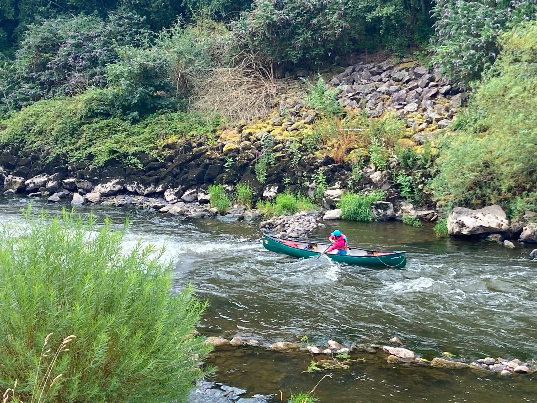 Person in a green canoe paddling down a rocky river surrounded by lush greenery.