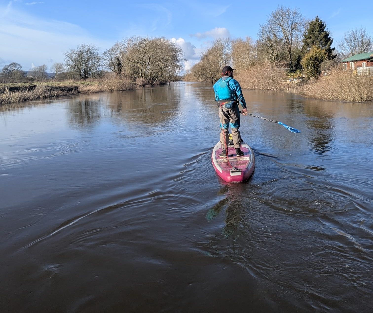 Person paddleboarding on a calm river under a clear blue sky.