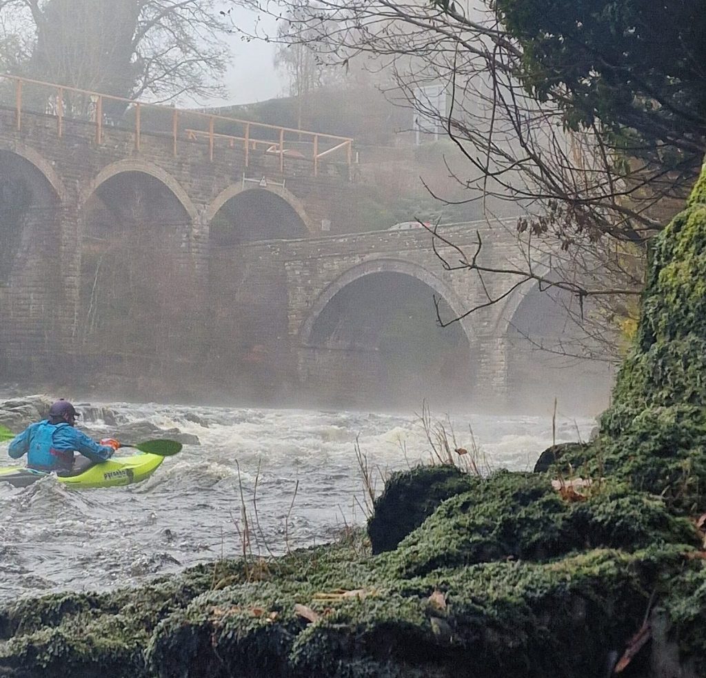 Kayaker on a misty river near an arched stone bridge.
