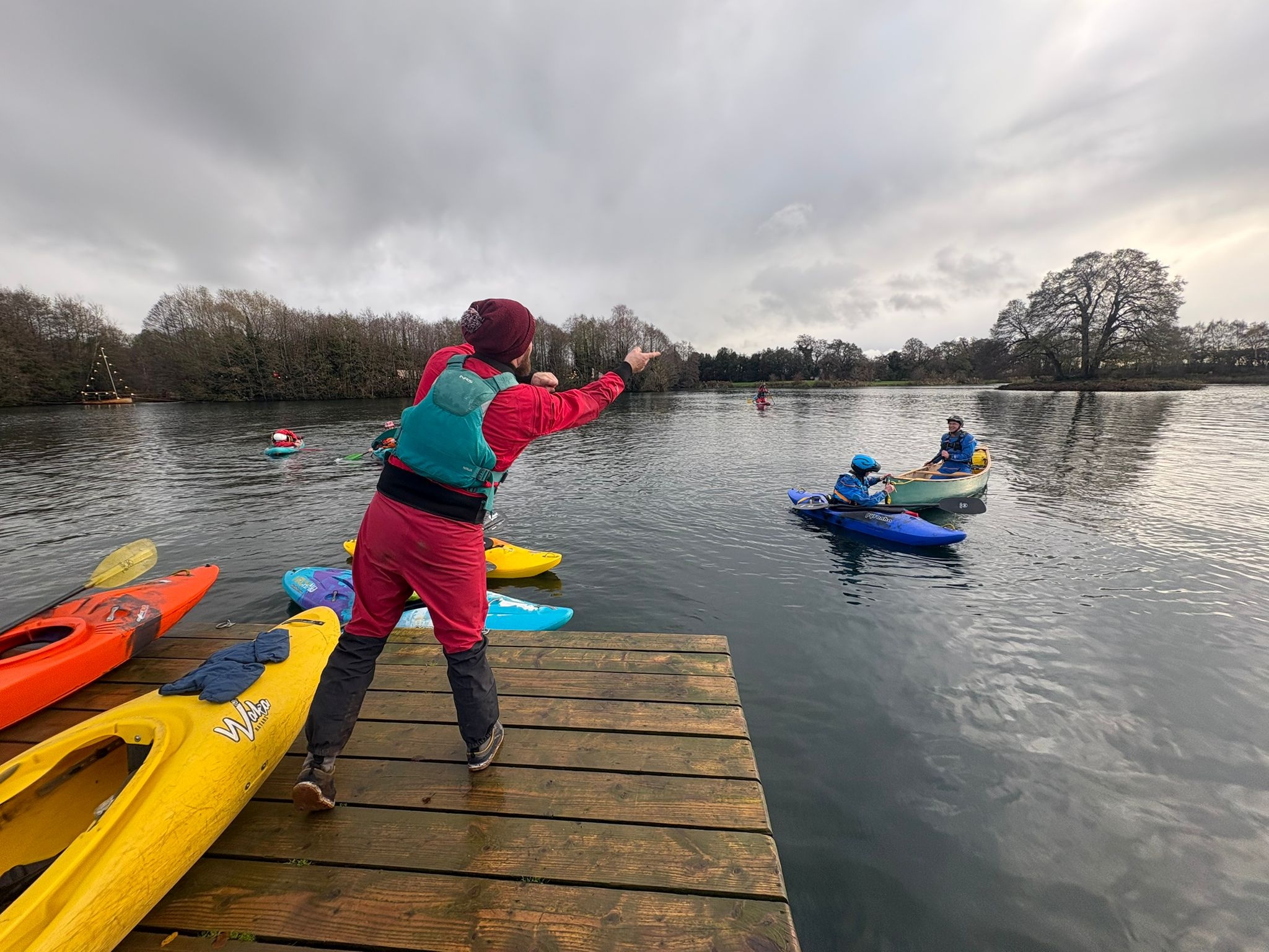 Person instructs kayakers on a lake from a wooden dock.
