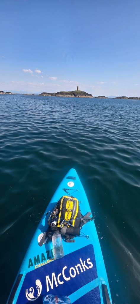 Blue paddleboard on calm sea facing distant island under clear sky.