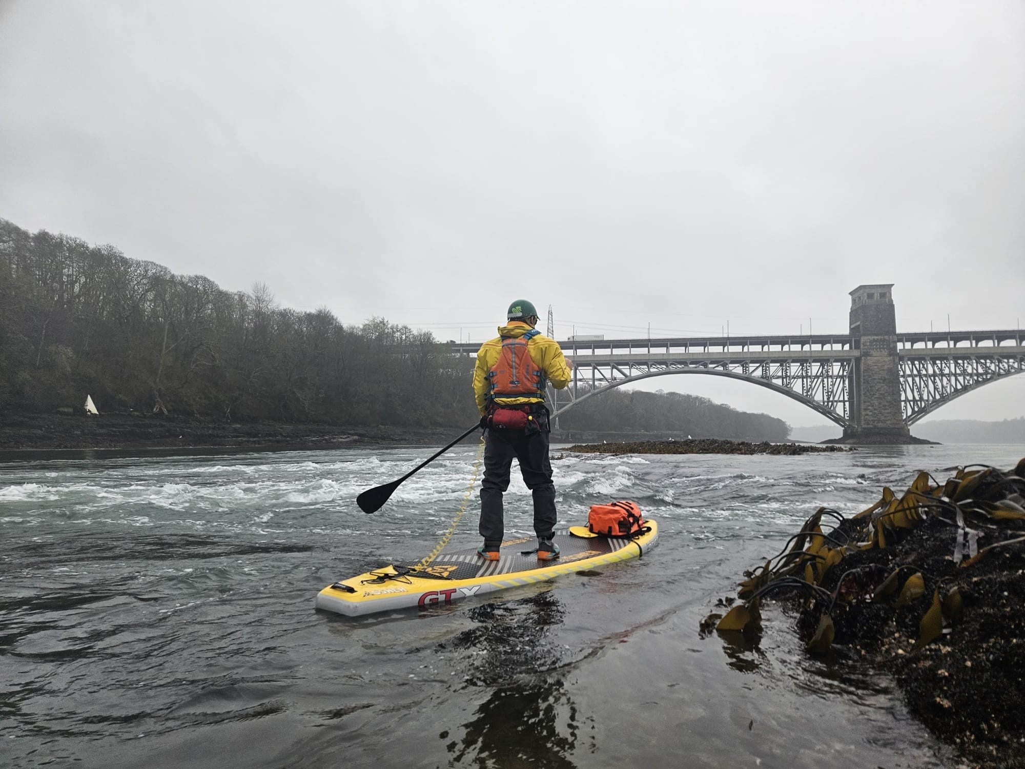 Person paddleboarding on river near bridge with overcast sky.