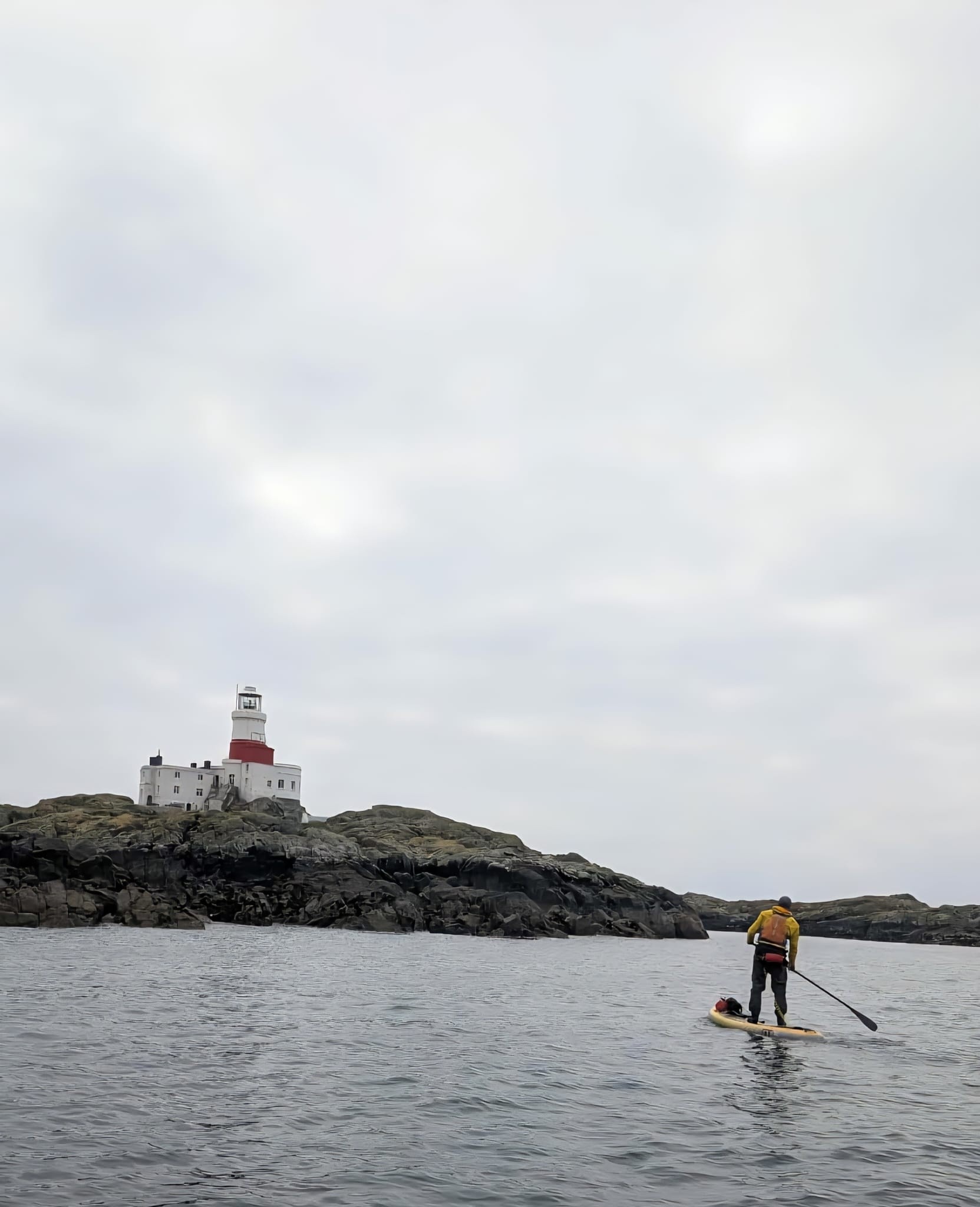 Person paddleboarding near a lighthouse on a rocky island.