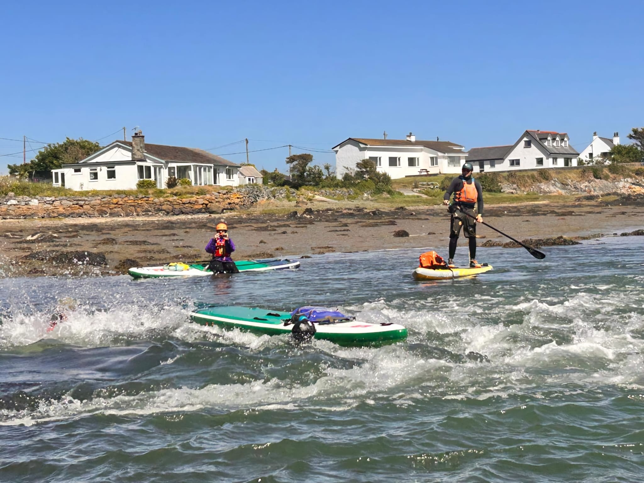People paddleboarding near a rocky shore with houses in the background.