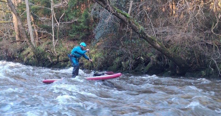 Person paddleboarding on a rough river with trees on the riverbank.