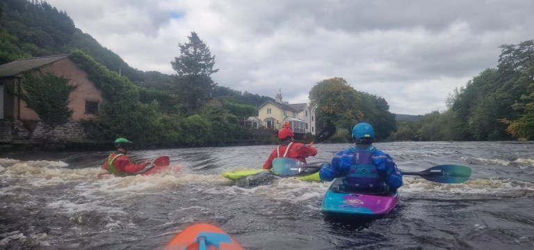 Three kayakers paddle on a river near a wooded shore and building.