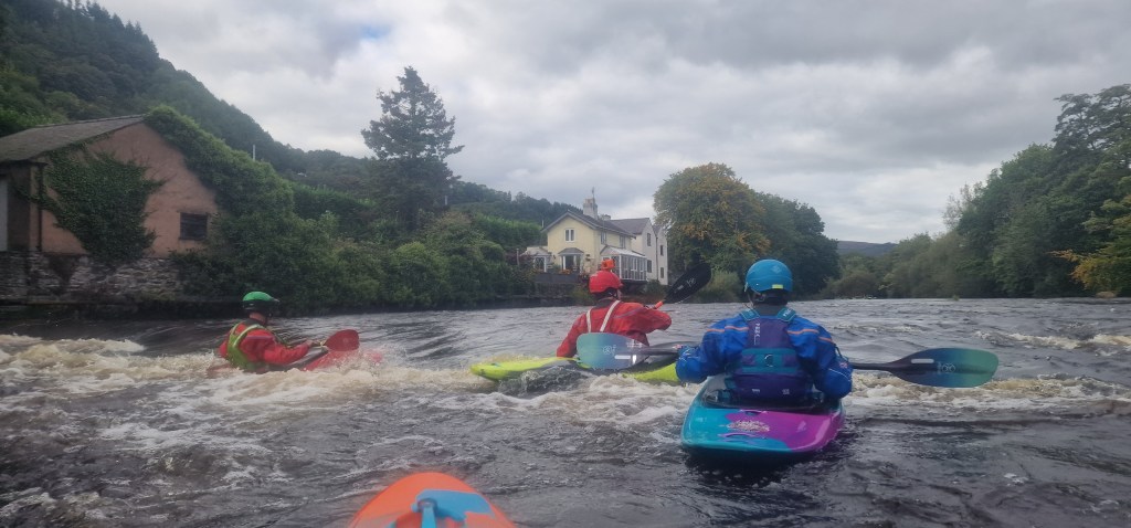 Three kayakers paddle on a river near a wooded shore and building.