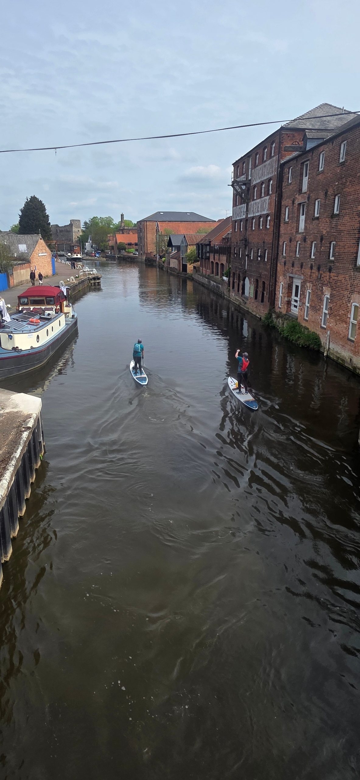 Paddleboarders on a river near brick buildings and moored boats.