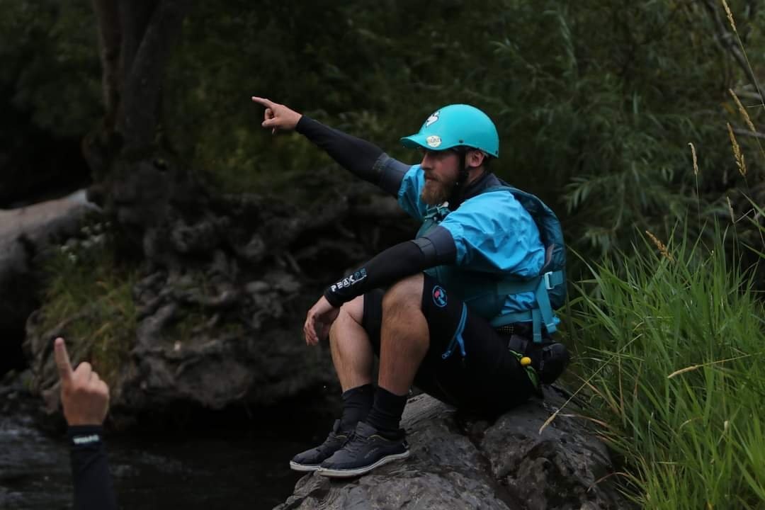 Person in blue gear sitting on rock, pointing forward near a stream.