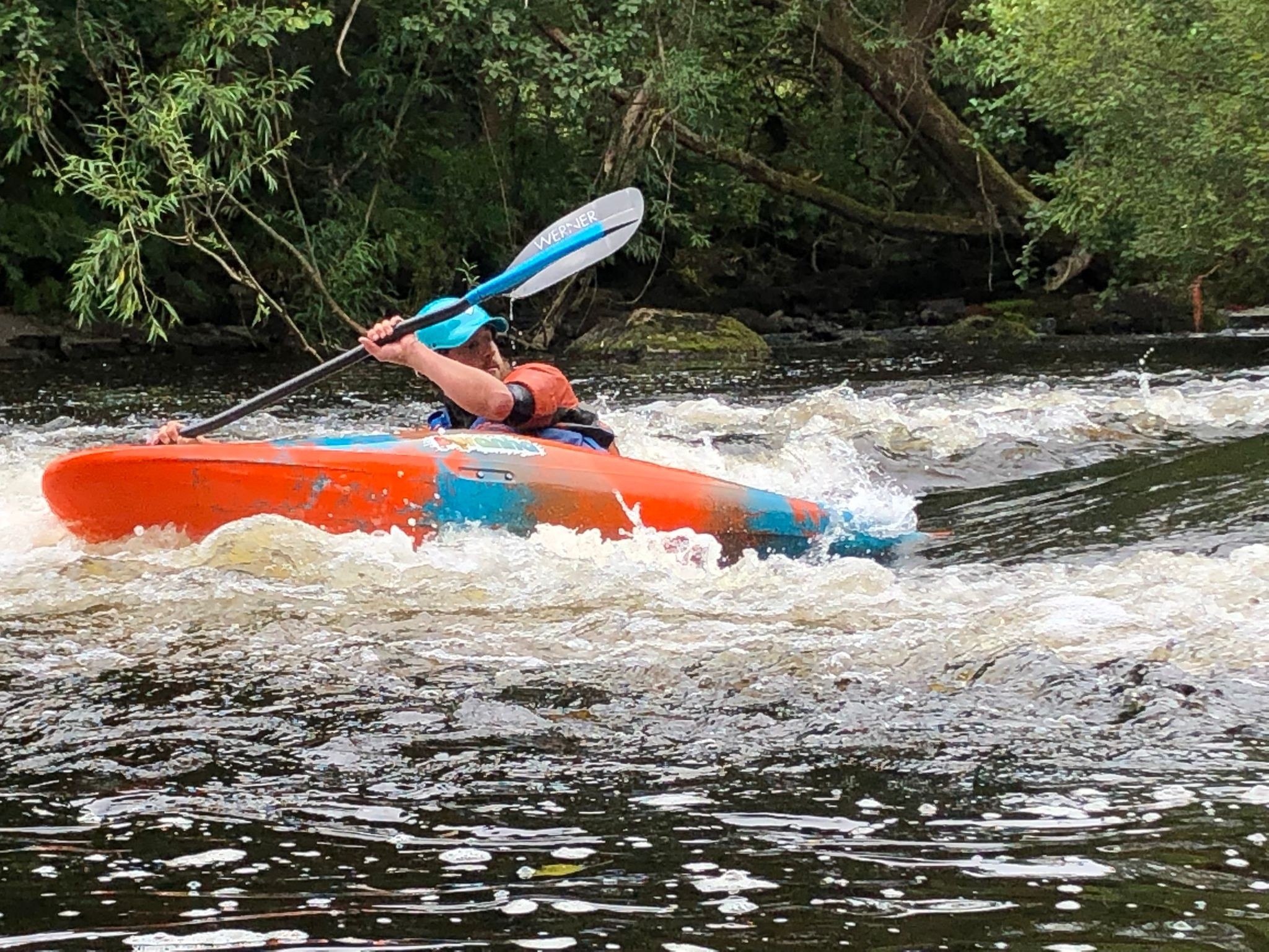 Person kayaking through whitewater in a river, surrounded by trees.
