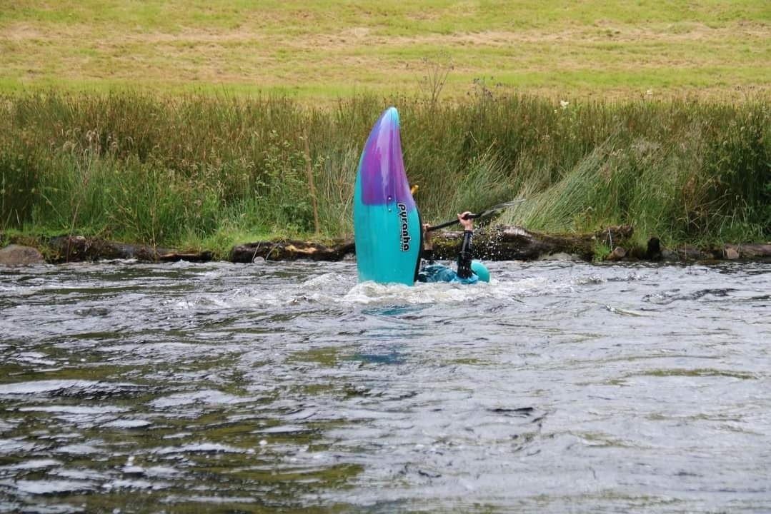 Capsized kayak with paddle submerged in a river near grassy bank.