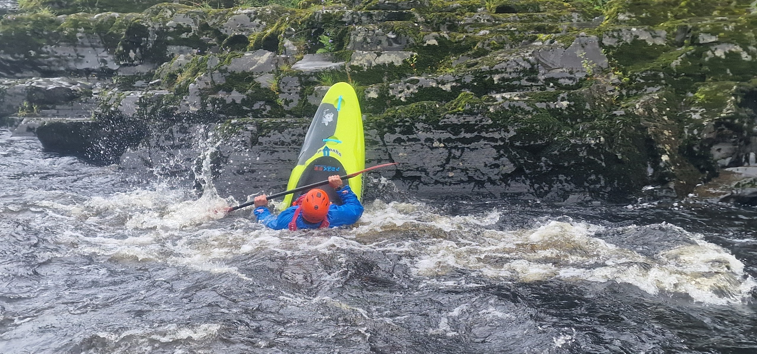 Person in a kayak capsizing in a river, near rocky riverbank.