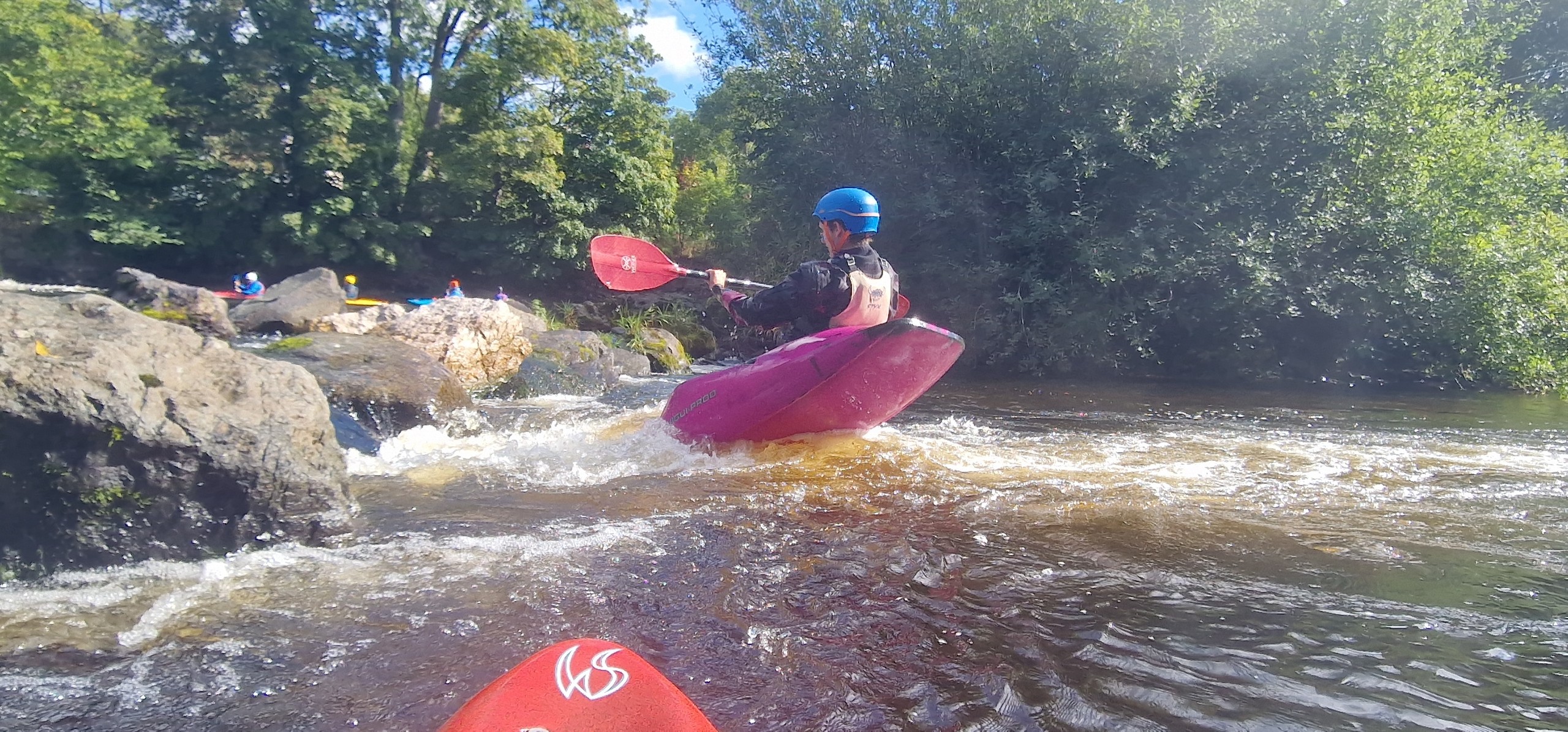 Person kayaking through a river with rocks and trees in the background.