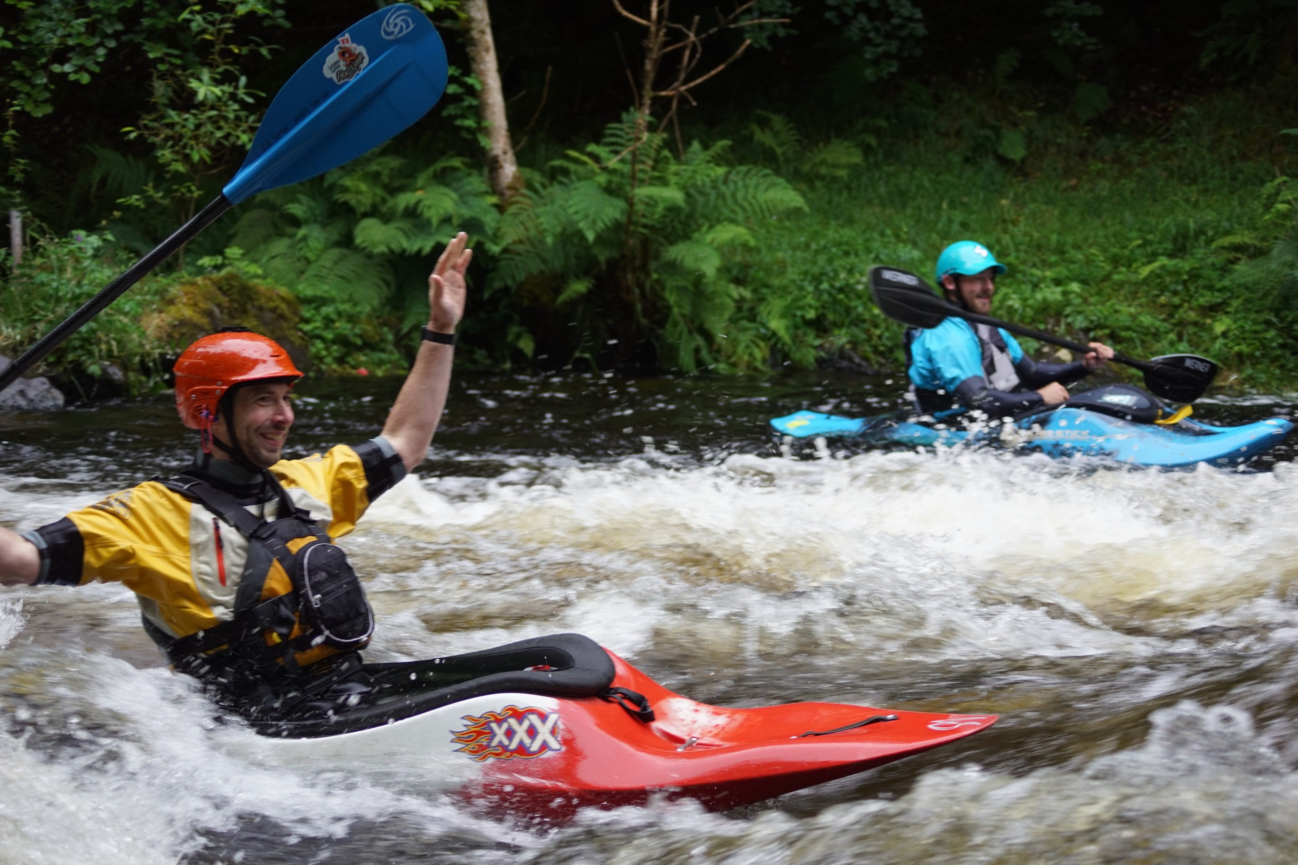 Two kayakers in helmets navigating whitewater rapids in a forested area.