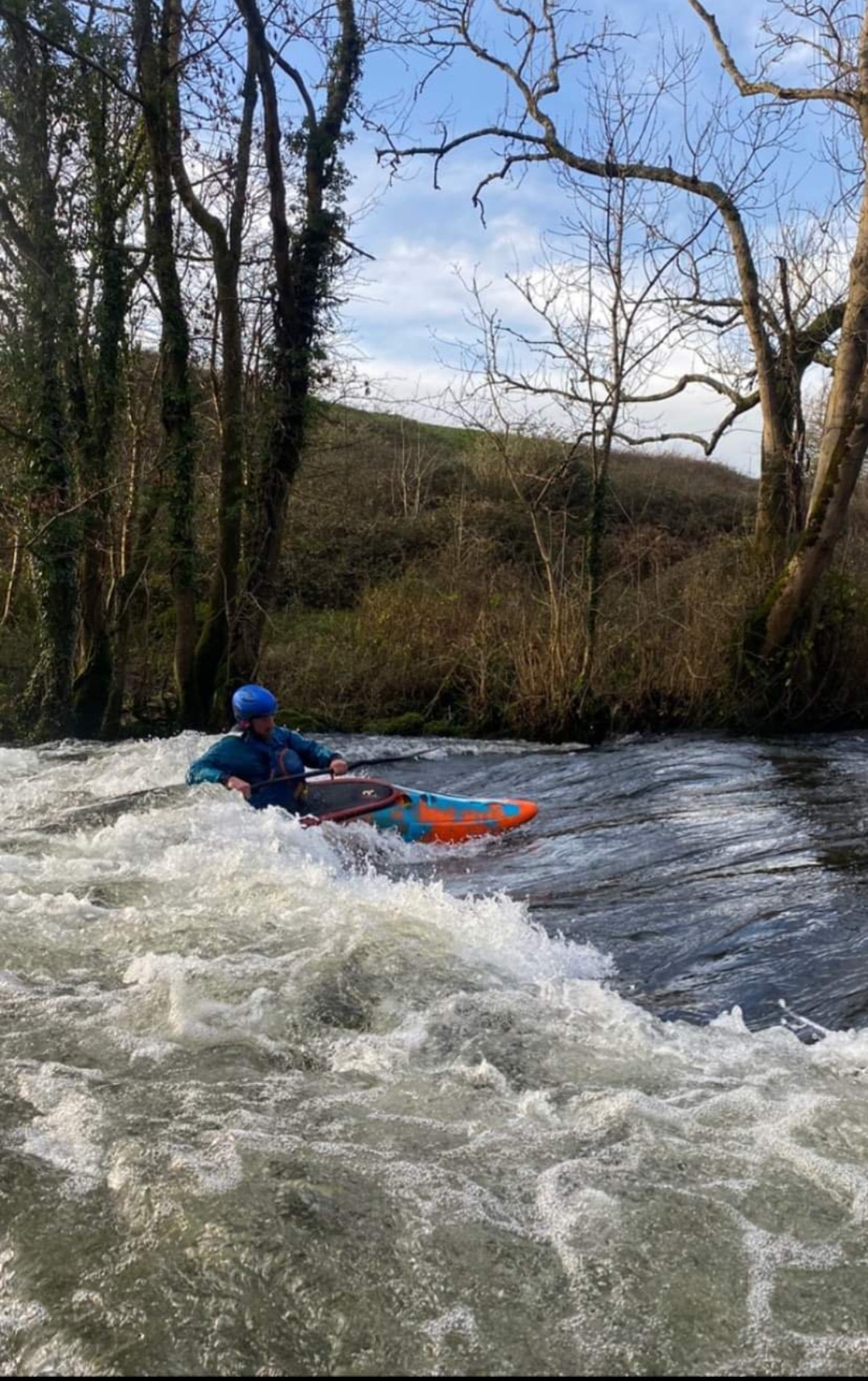Person kayaking through rapids with trees around and a cloudy sky.