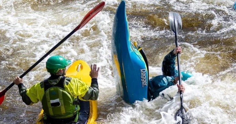 Kayaker capsizes in whitewater, another kayaker nearby offers assistance.
