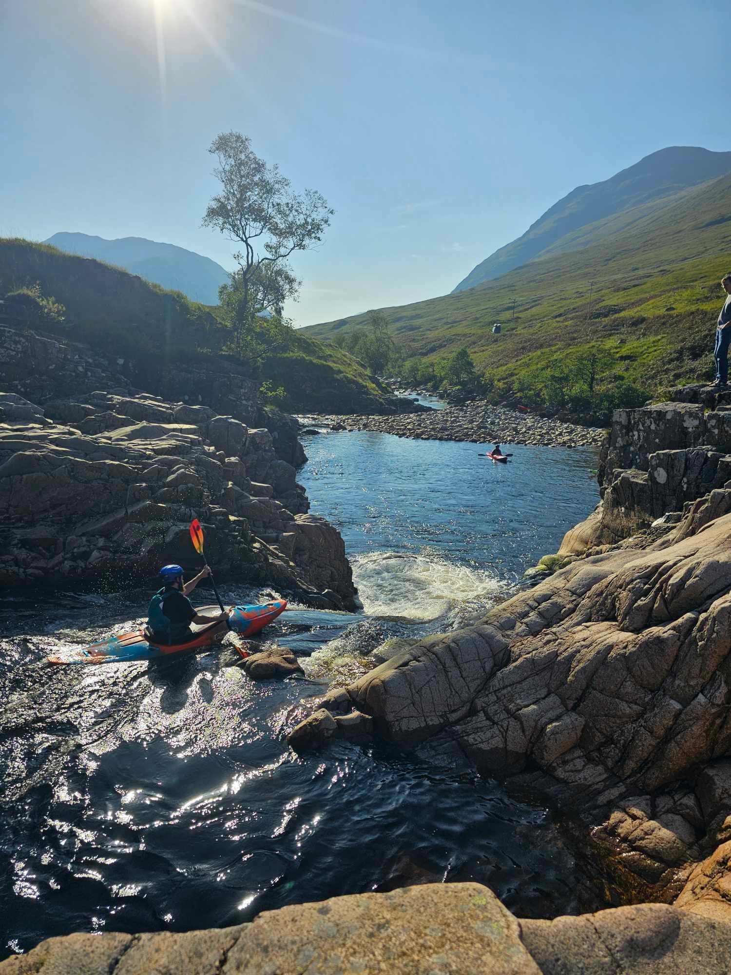 Kayaker paddling in a rocky river under bright sun, with mountains in the background.