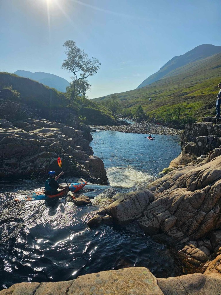 Kayaker paddling in a rocky river under bright sun, with mountains in the background.