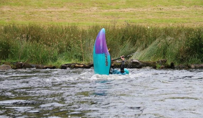 Kayaker vertically balancing in river near grassy bank.