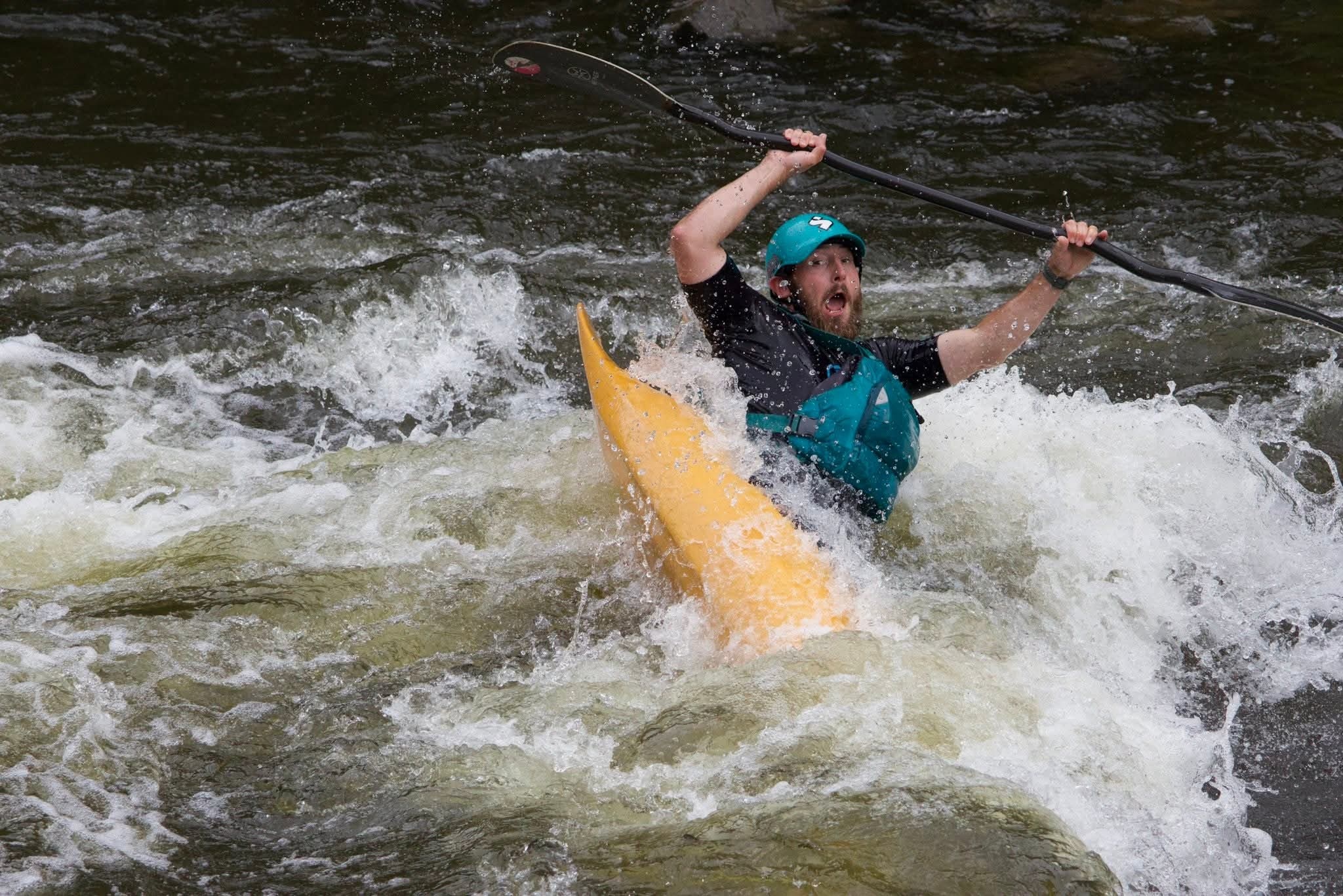 Person kayaking through whitewater rapids with a paddle raised.