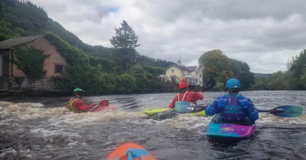 Kayakers paddling on a river near houses and trees under a cloudy sky.
