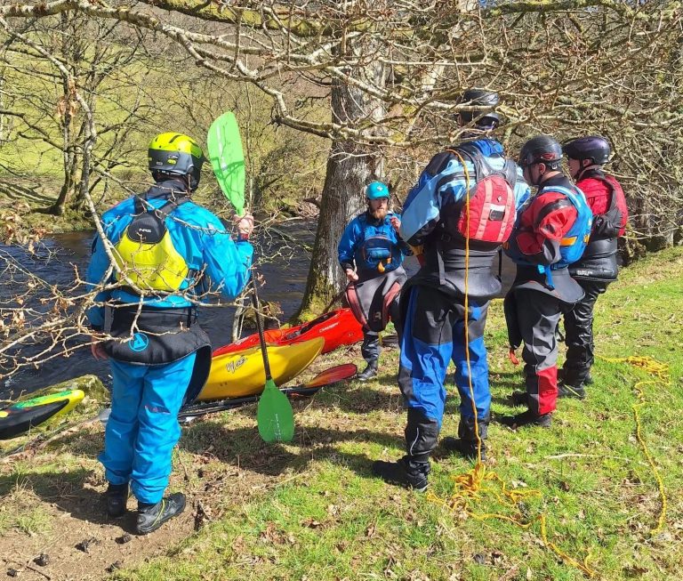 Group of kayakers in gear stand by river with trees.