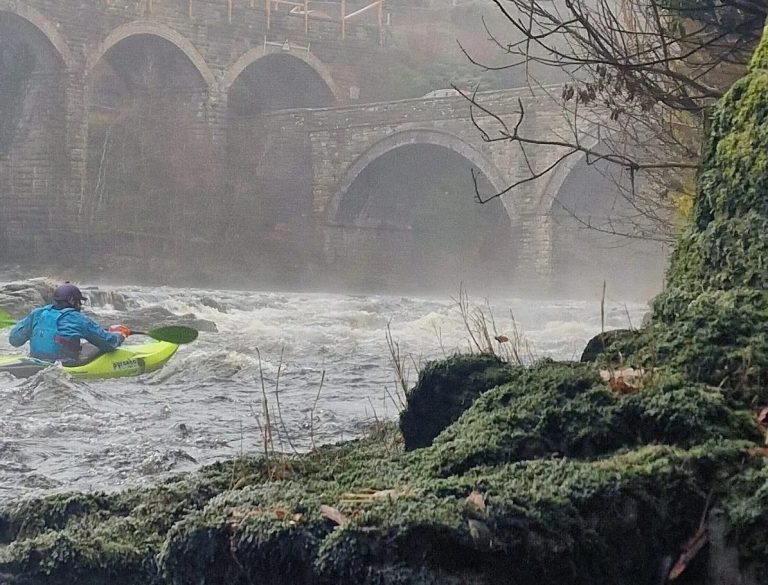 Person kayaking on a river near old arched bridge in misty weather.