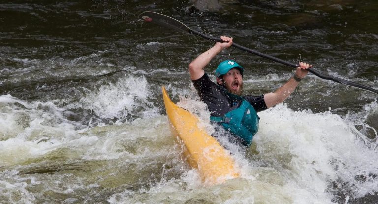 Person kayaking through whitewater rapids, wearing a teal helmet and vest.