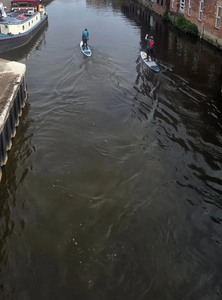 Two people paddleboarding on a calm canal surrounded by buildings.