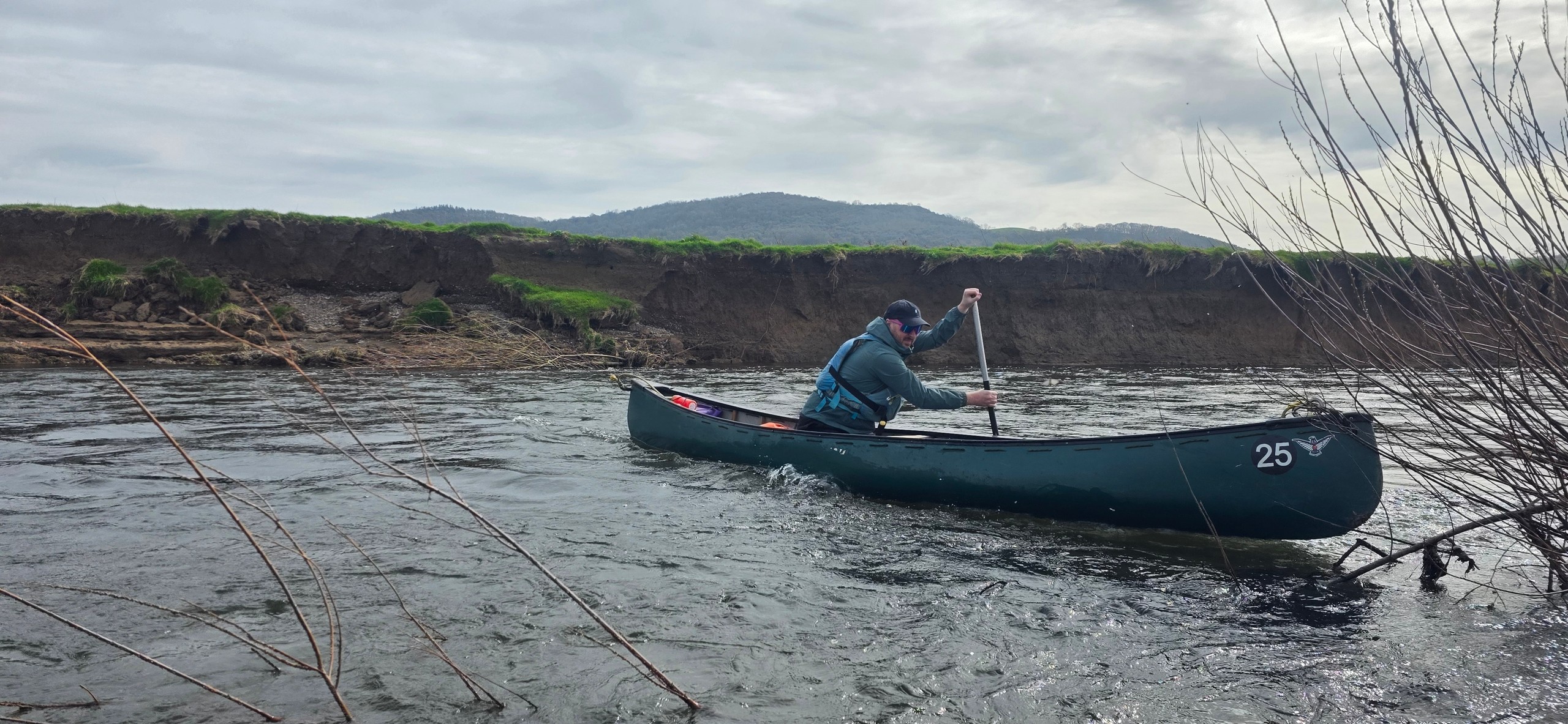 Person canoeing on a river with grassy banks and hills in the background.