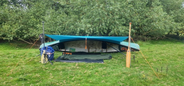 Inverted canoe with tarp shelter on grassy field near trees.