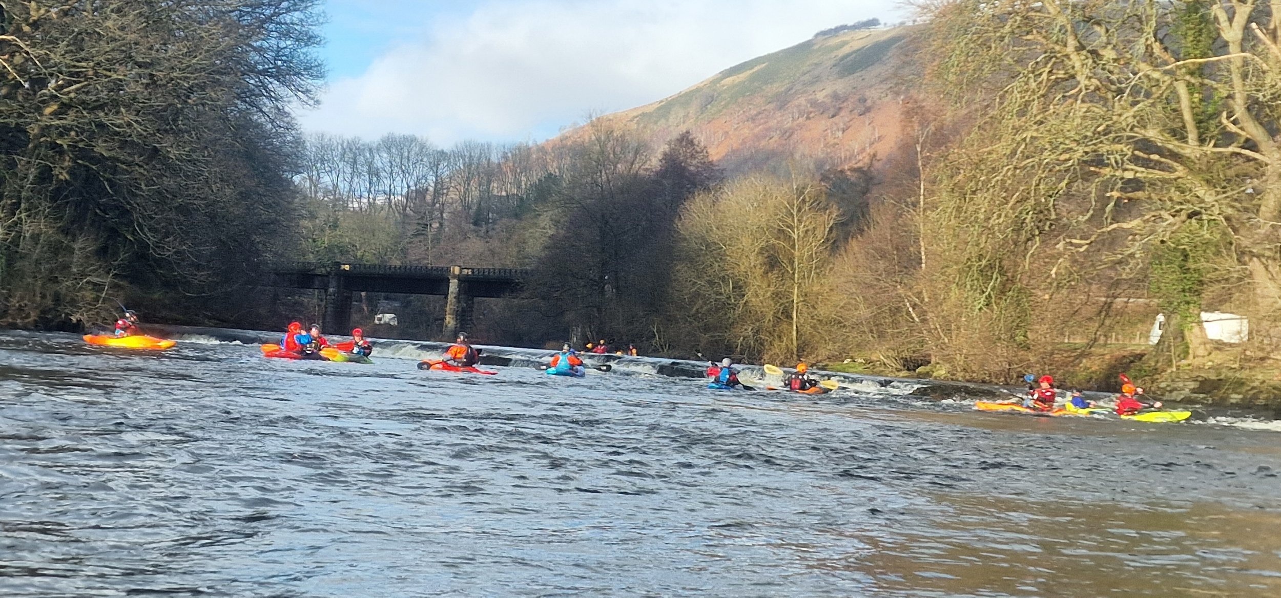 Group of kayakers paddling on a river surrounded by trees and hills.