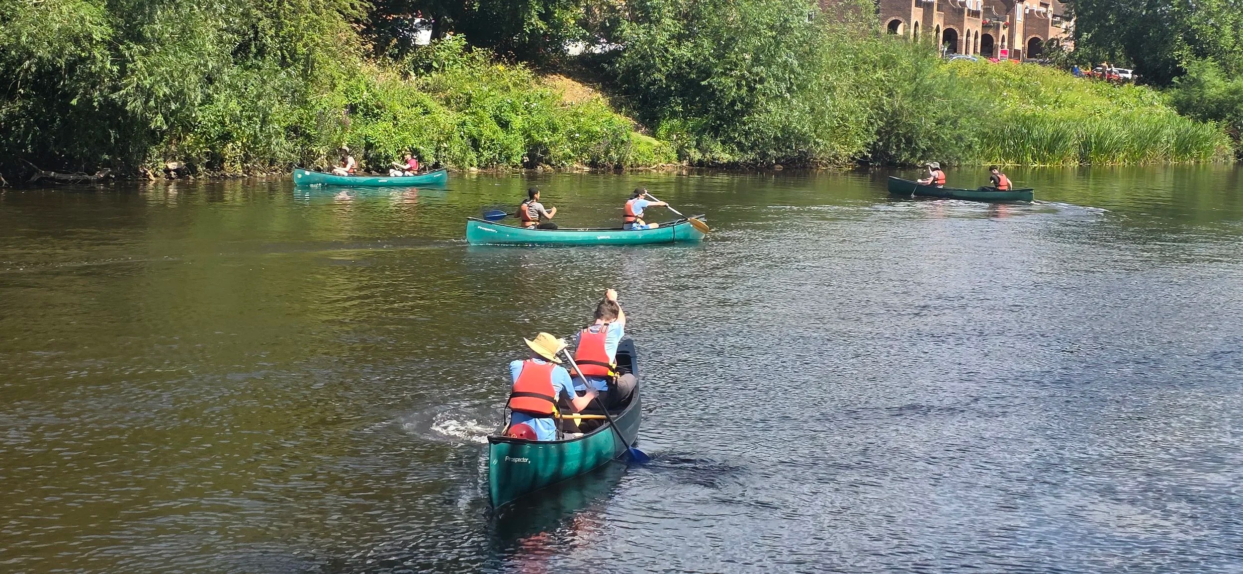 People canoeing on a river surrounded by greenery and buildings in the background.