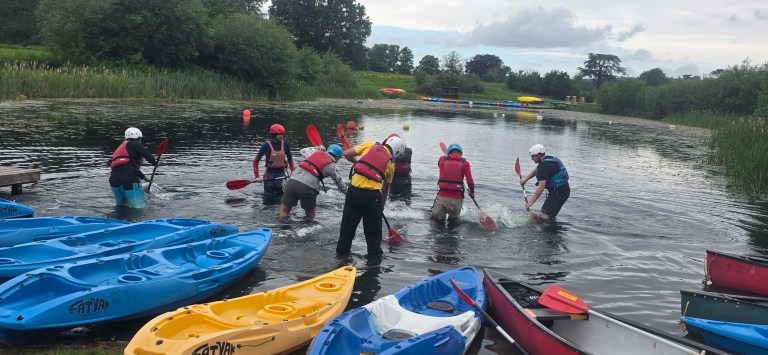 People in life vests and helmets playing in a small lake with kayaks nearby.